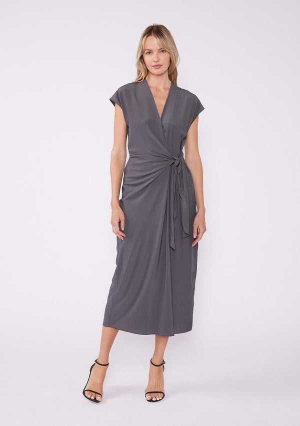 [Color: Pewter] A front facing image of a blonde model wearing a dark grey pewter maxi length wrap dress. With short cap sleeves, a deep v neckline, and a side tie waist closure. A beautiful office dress.