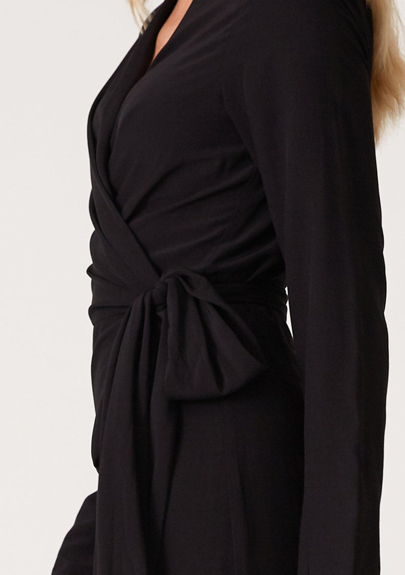 [Color: Black] A close up side facing image of a blonde model wearing a sophisticated black mid length wrap dress with long sleeves, a v neckline, a side slit, and a side tie waist. 