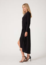 [Color: Black] A side facing image of a blonde model wearing a sophisticated black mid length wrap dress with long sleeves, a v neckline, a side slit, and a side tie waist. 