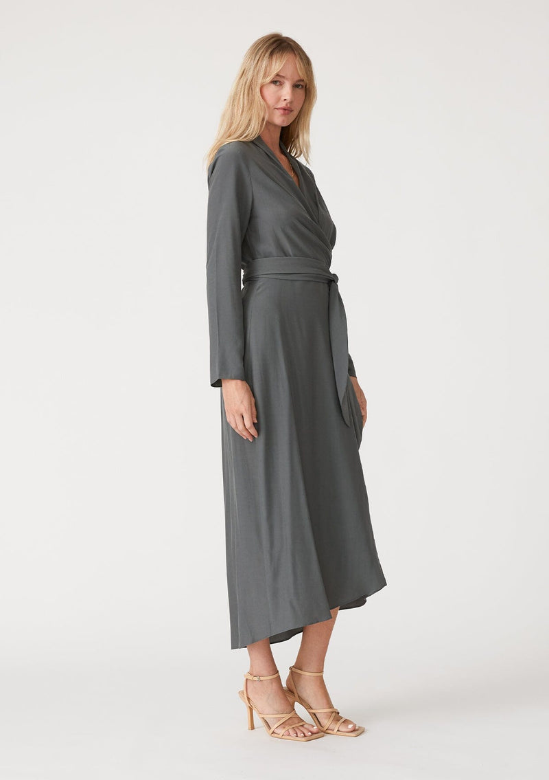 [Color: Moss] A side facing image of a blonde model wearing a sophisticated moss green mid length wrap dress with long sleeves, a v neckline, a side slit, and a side tie waist.