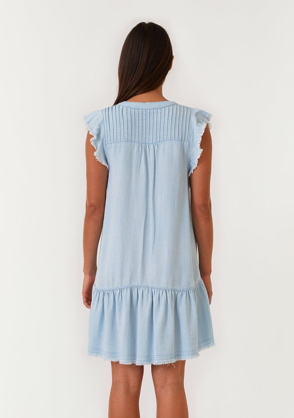 [Color: Bleach Wash] A back facing image of a brunette model wearing a denim blue bohemian spring mini dress crafted from Tencel. With short flutter sleeves, a v neckline, a tiered mini skirt, front patch pockets, a self covered button front, a raw hemline, and pleated details. 