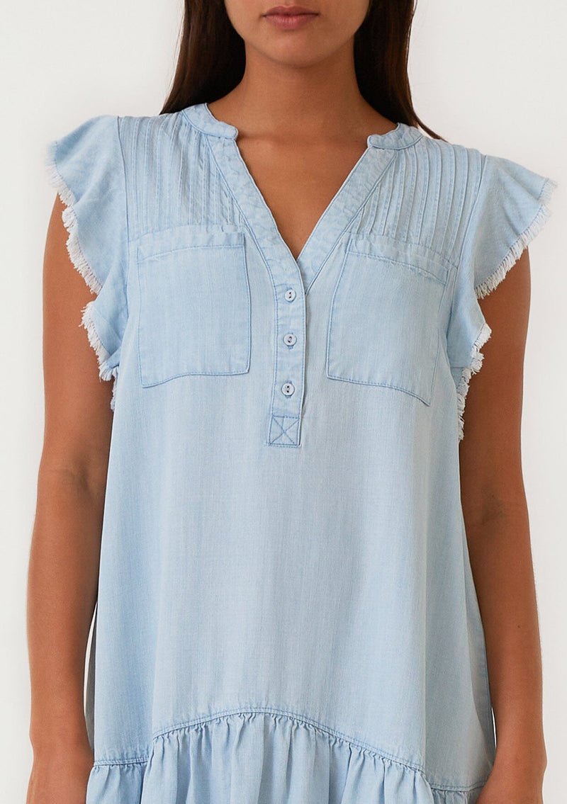 [Color: Bleach Wash] A close up front facing image of a brunette model wearing a denim blue bohemian spring mini dress crafted from Tencel. With short flutter sleeves, a v neckline, a tiered mini skirt, front patch pockets, a self covered button front, a raw hemline, and pleated details. 