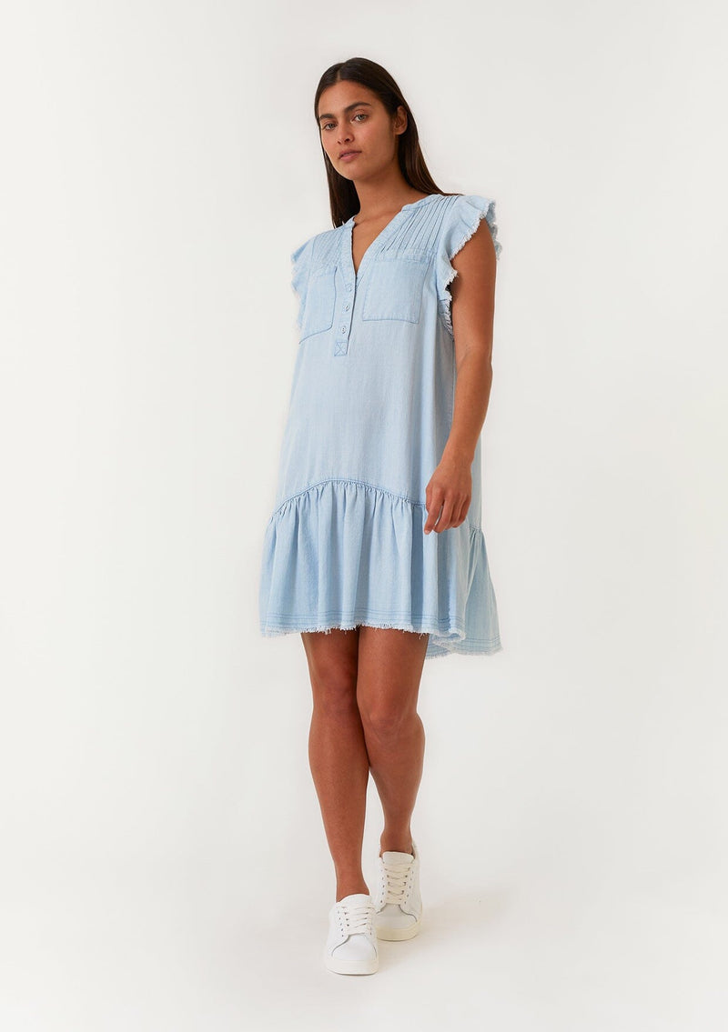 [Color: Bleach Wash] A full body front facing image of a brunette model wearing a denim blue bohemian spring mini dress crafted from Tencel. With short flutter sleeves, a v neckline, a tiered mini skirt, front patch pockets, a self covered button front, a raw hemline, and pleated details. 