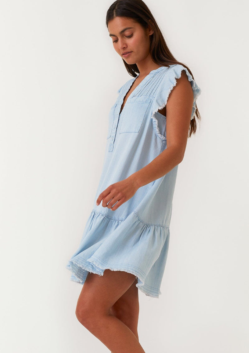 [Color: Bleach Wash] A side facing image of a brunette model wearing a denim blue bohemian spring mini dress crafted from Tencel. With short flutter sleeves, a v neckline, a tiered mini skirt, front patch pockets, a self covered button front, a raw hemline, and pleated details. 