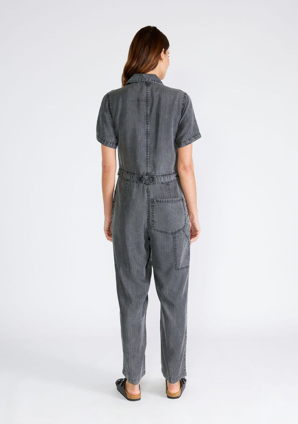 [Color: Ash Grey Wash] A back facing image of a brunette model wearing a utility style jumpsuit in an ash grey wash. Designed in Tencel, with short sleeves, a collared neckline, a long straight leg, a button front top, a zip fly closure, and front and back patch pockets. 