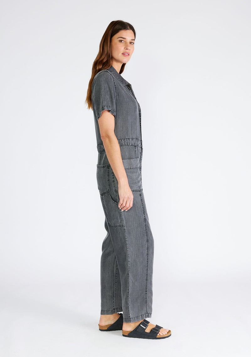 [Color: Ash Grey Wash] A side facing image of a brunette model wearing a utility style jumpsuit in an ash grey wash. Designed in Tencel, with short sleeves, a collared neckline, a long straight leg, a button front top, a zip fly closure, and front and back patch pockets. 