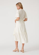 [Color: Vanilla] A back facing image of a blonde model wearing an off white bohemian mid length wrap dress. With short flutter sleeves, embroidered detail, a deep v neckline, a side tie waist closure, and a flowy tiered skirt. 