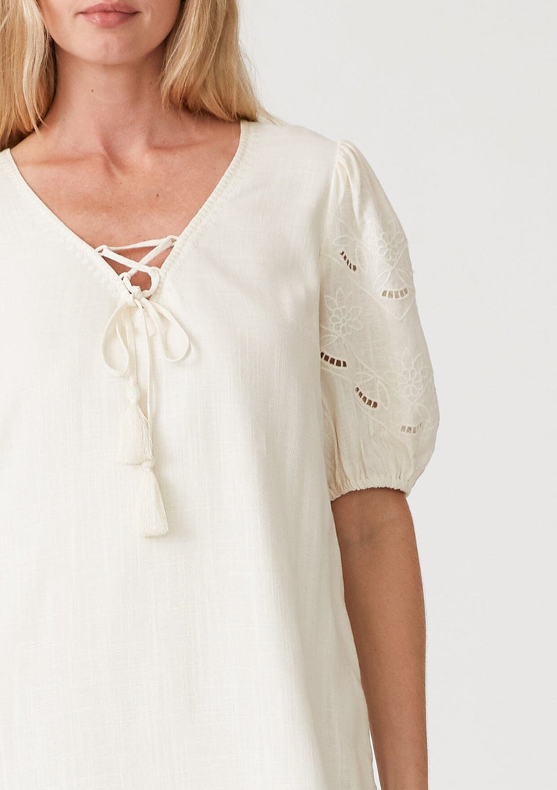 [Color: Vanilla] A close up front facing image of a blonde model wearing an off white bohemian mini dress with short puff sleeves, embroidered detail, side pockets, a v neckline with lace up detail and tassel ties, and a relaxed, loose fit. 