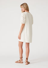 [Color: Vanilla] A back facing image of a blonde model wearing an off white bohemian mini dress with short puff sleeves, embroidered detail, side pockets, a v neckline with lace up detail and tassel ties, and a relaxed, loose fit. 