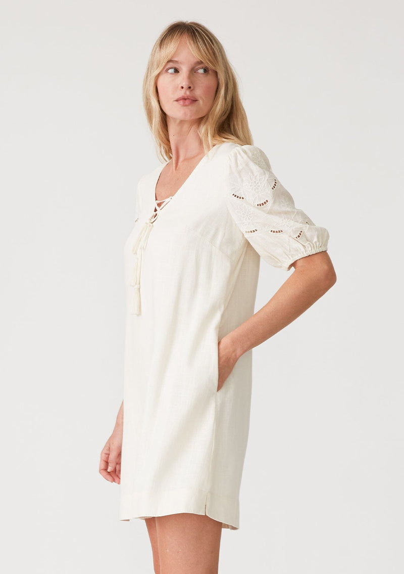 [Color: Vanilla] A side facing image of a blonde model wearing an off white bohemian mini dress with short puff sleeves, embroidered detail, side pockets, a v neckline with lace up detail and tassel ties, and a relaxed, loose fit. 