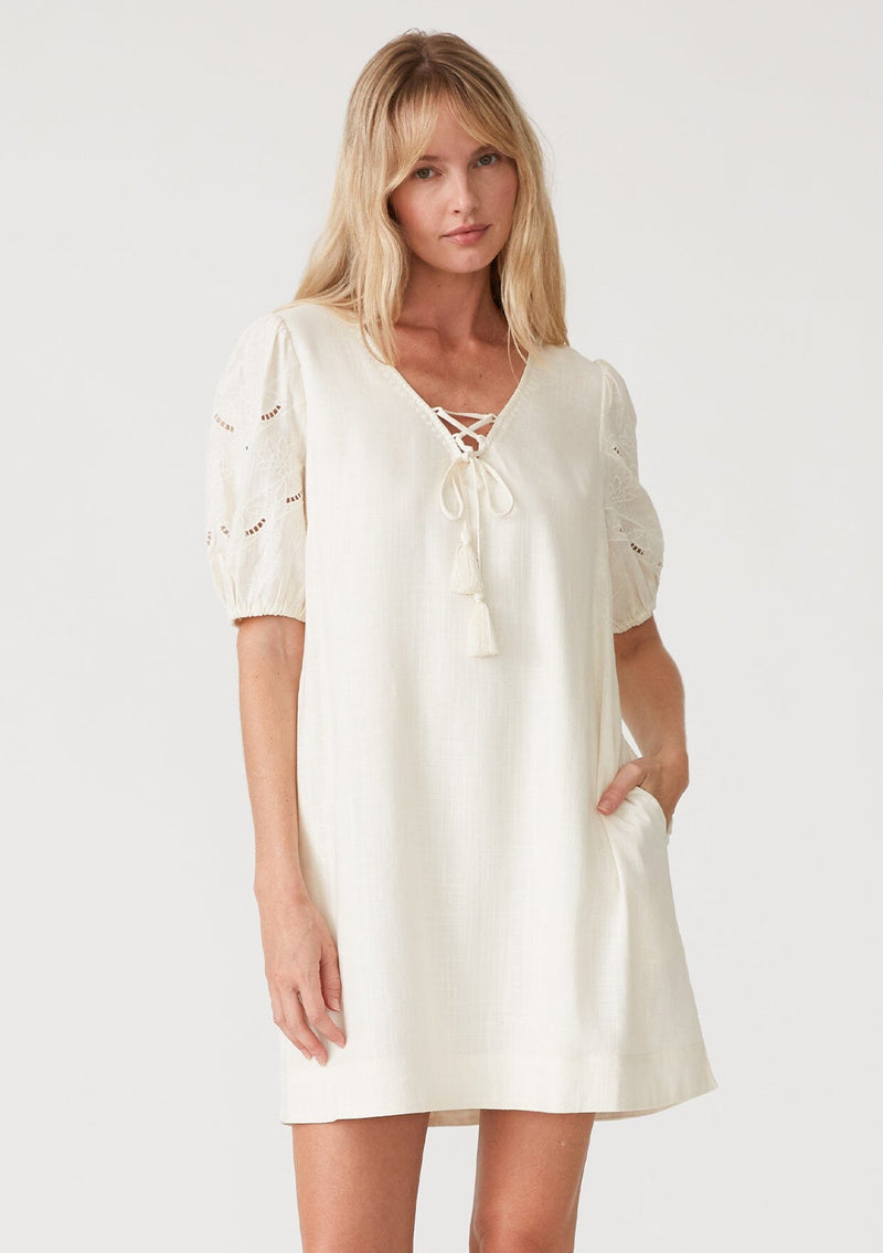 [Color: Vanilla] A front facing image of a blonde model wearing an off white bohemian mini dress with short puff sleeves, embroidered detail, side pockets, a v neckline with lace up detail and tassel ties, and a relaxed, loose fit. 