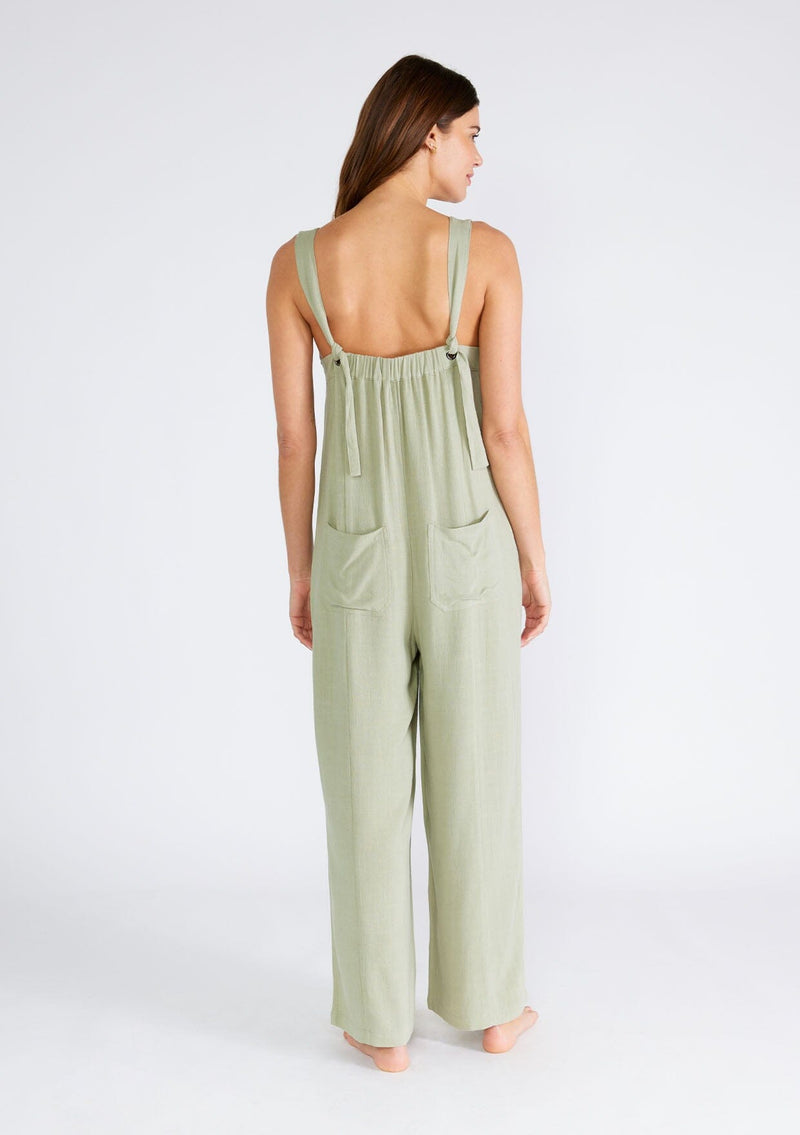 [Color: Dusty Sage] A back facing image of a brunette model wearing a bohemian sleeveless jumpsuit in a dusty sage green. With adjustable tank top straps, a v neckline, a long wide leg, side pockets, and back patch pockets. 