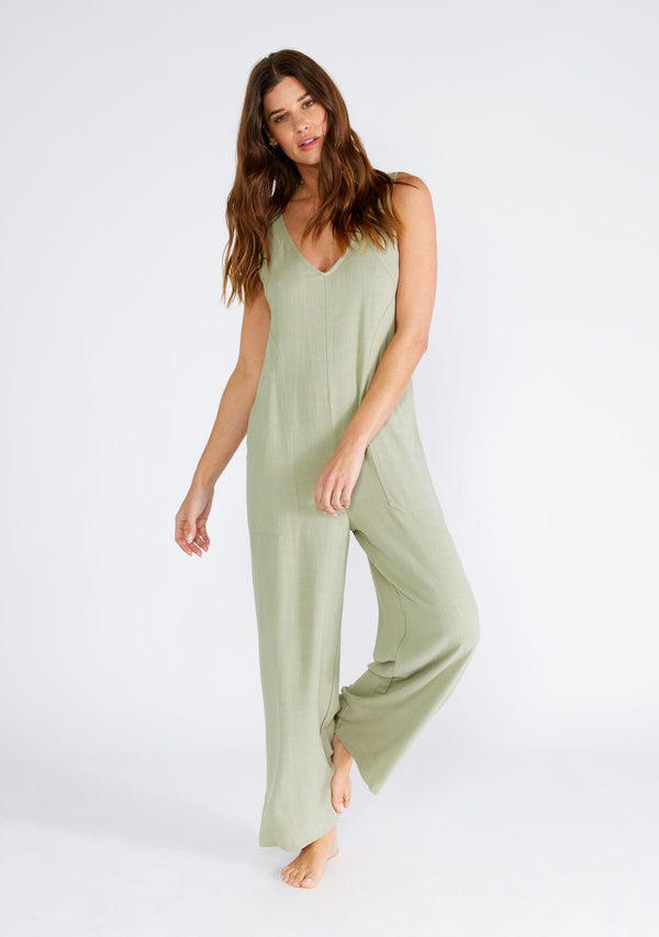 [Color: Dusty Sage] A front facing image of a brunette model wearing a bohemian sleeveless jumpsuit in a dusty sage green. With adjustable tank top straps, a v neckline, a long wide leg, side pockets, and back patch pockets. 