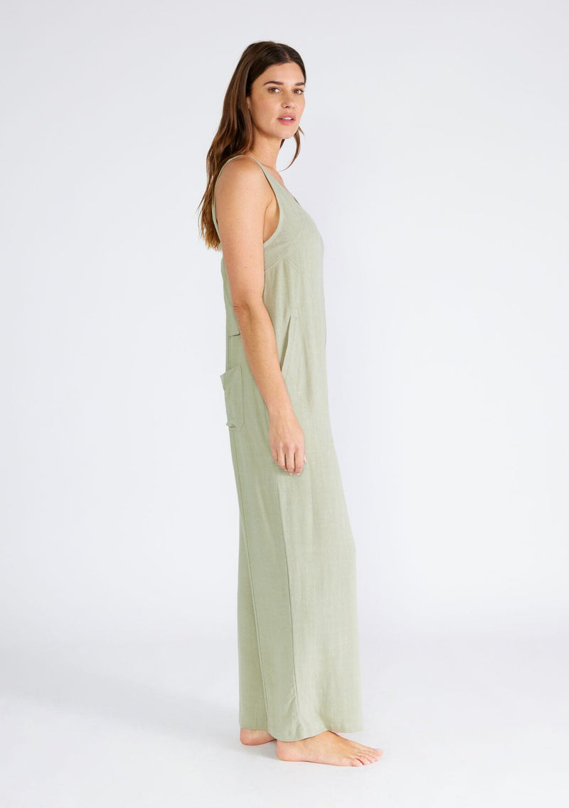 [Color: Dusty Sage] A side facing image of a brunette model wearing a bohemian sleeveless jumpsuit in a dusty sage green. With adjustable tank top straps, a v neckline, a long wide leg, side pockets, and back patch pockets. 