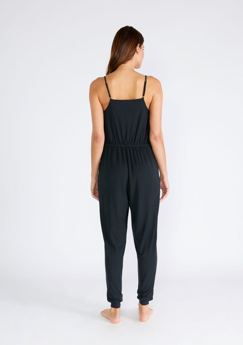 [Color: Black] A back facing image of a brunette model wearing a black ribbed knit sleeveless jumpsuit. With spaghetti straps, a surplice v neckline, side pockets, and a drawstring tie waist. 