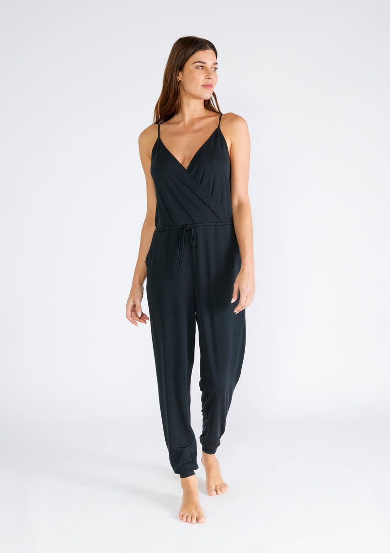[Color: Black] A full body front facing image of a brunette model wearing a black ribbed knit sleeveless jumpsuit. With spaghetti straps, a surplice v neckline, side pockets, and a drawstring tie waist. 