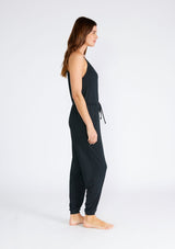 [Color: Black] A side facing image of a brunette model wearing a black ribbed knit sleeveless jumpsuit. With spaghetti straps, a surplice v neckline, side pockets, and a drawstring tie waist. 