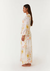 [Color: Vanilla/Coral] A side facing image of a brunette model wearing a dreamy bohemian maxi dress designed in an ivory and coral pink floral print. With long bell sleeves, a v neckline, a self covered button front, a long flowy skirt with front slit, lace trim, and an adjustable drawstring waist with tassel ties. 