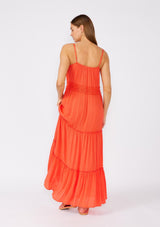 [Color: Coral] A back facing image of a brunette model wearing a bright coral red sleeveless maxi dress. With adjustable spaghetti straps, a straight neckline, crochet trim, a tassel tie drawstring waist, and a ruffle trimmed tiered skirt. 