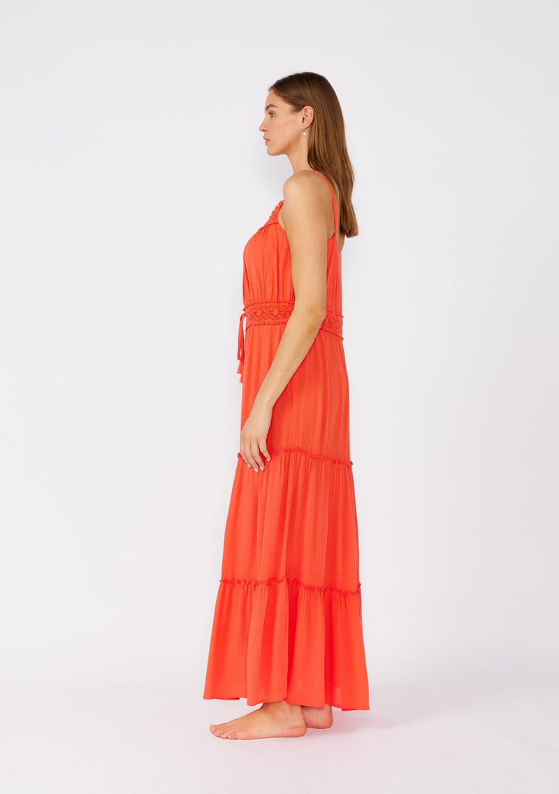 [Color: Coral] A side facing image of a brunette model wearing a bright coral red sleeveless maxi dress. With adjustable spaghetti straps, a straight neckline, crochet trim, a tassel tie drawstring waist, and a ruffle trimmed tiered skirt. 