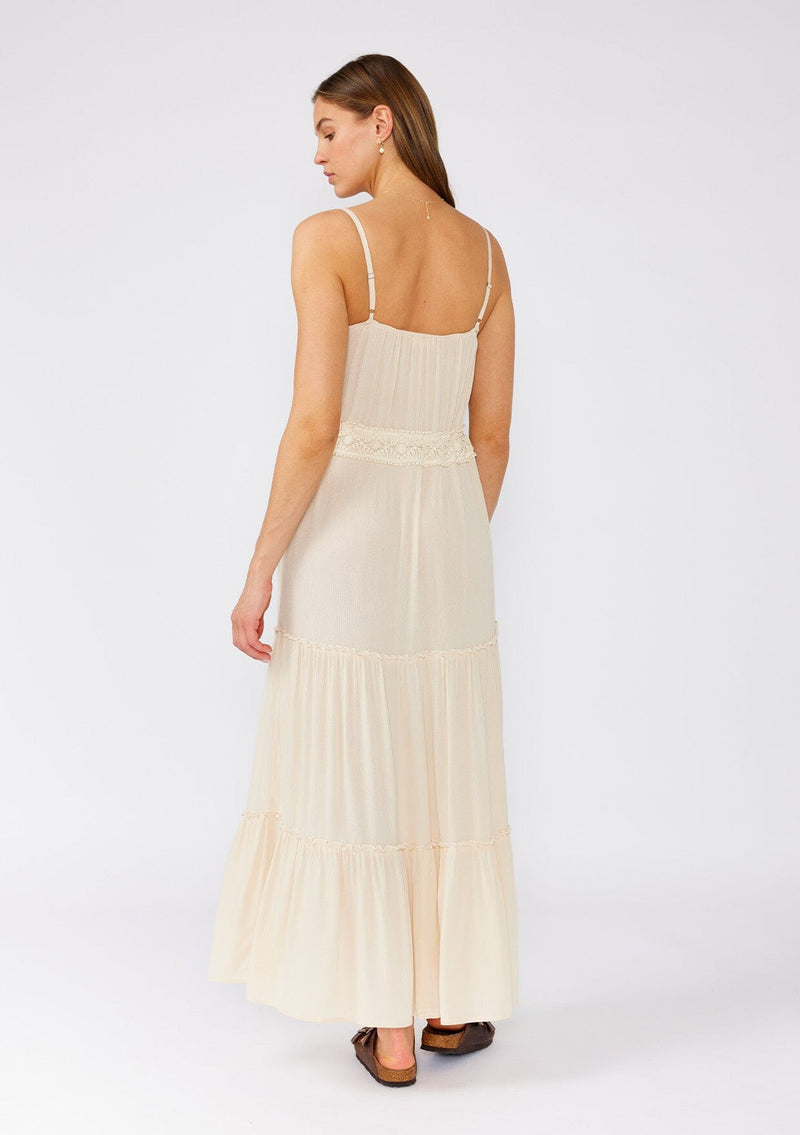 [Color: Natural] A back facing image of a brunette model wearing an off white sleeveless maxi dress. With adjustable spaghetti straps, a straight neckline, crochet trim, a tassel tie drawstring waist, and a ruffle trimmed tiered skirt. 