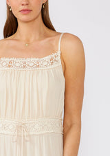 [Color: Natural] A close up front facing image of a brunette model wearing an off white sleeveless maxi dress. With adjustable spaghetti straps, a straight neckline, crochet trim, a tassel tie drawstring waist, and a ruffle trimmed tiered skirt. 