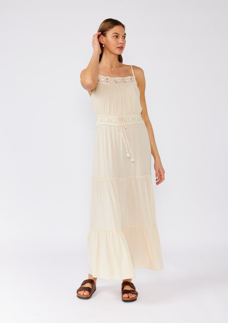 [Color: Natural] A front facing image of a brunette model wearing an off white sleeveless maxi dress. With adjustable spaghetti straps, a straight neckline, crochet trim, a tassel tie drawstring waist, and a ruffle trimmed tiered skirt. 