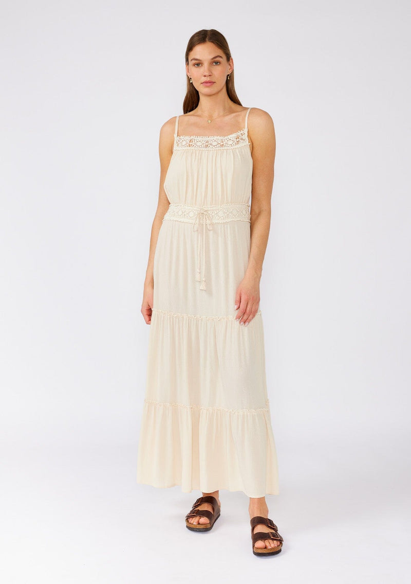 [Color: Natural] A full body front facing image of a brunette model wearing an off white sleeveless maxi dress. With adjustable spaghetti straps, a straight neckline, crochet trim, a tassel tie drawstring waist, and a ruffle trimmed tiered skirt. 