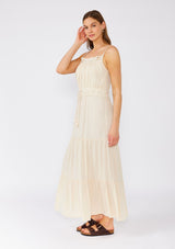 [Color: Natural] A side facing image of a brunette model wearing an off white sleeveless maxi dress. With adjustable spaghetti straps, a straight neckline, crochet trim, a tassel tie drawstring waist, and a ruffle trimmed tiered skirt. 