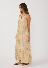 [Color: Dusty Peach/Rust] A side facing image of a brunette model wearing a bohemian sleeveless maxi dress designed in a peach and rust floral print. With a collared neckline, a self covered button front top, a long tiered skirt, and side pockets. 