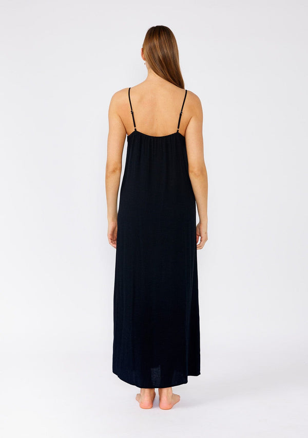 [Color: Black] A back facing image of a brunette model wearing a classic black maxi slip dress with lace detail along the neckline. With adjustable spaghetti straps, a scooped neckline, and a long flowy skirt with side slits. 