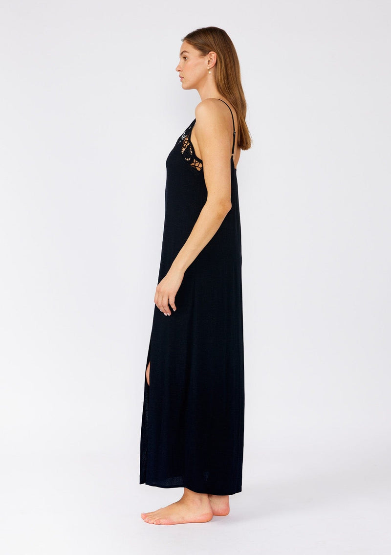 [Color: Black] A side facing image of a brunette model wearing a classic black maxi slip dress with lace detail along the neckline. With adjustable spaghetti straps, a scooped neckline, and a long flowy skirt with side slits. 