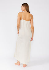 [Color: Natural] A back facing image of a brunette model wearing a classic off white maxi slip dress with lace detail along the neckline. With adjustable spaghetti straps, a scooped neckline, and a long flowy skirt with side slits. 