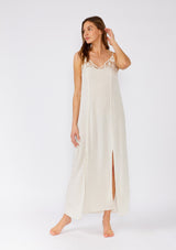 [Color: Natural] A front facing image of a brunette model wearing a classic off white maxi slip dress with lace detail along the neckline. With adjustable spaghetti straps, a scooped neckline, and a long flowy skirt with side slits. 