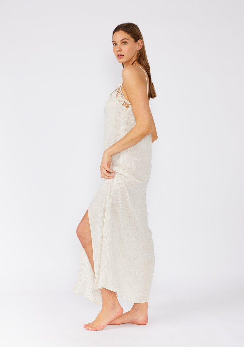 [Color: Natural] A side facing image of a brunette model wearing a classic off white maxi slip dress with lace detail along the neckline. With adjustable spaghetti straps, a scooped neckline, and a long flowy skirt with side slits. 