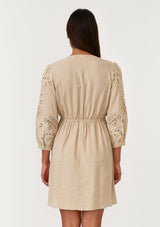 [Color: Natural] A back facing image of a brunette model wearing a classic bohemian natural brown mini dress in a linen blend. With three quarter length sleeves, multi colored embroidered detail, a v neckline, an empire waist, and a relaxed fit. 