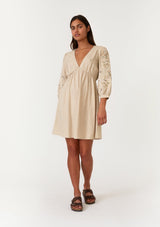 [Color: Natural] A full body front facing image of a brunette model wearing a classic bohemian natural brown mini dress in a linen blend. With three quarter length sleeves, multi colored embroidered detail, a v neckline, an empire waist, and a relaxed fit. 