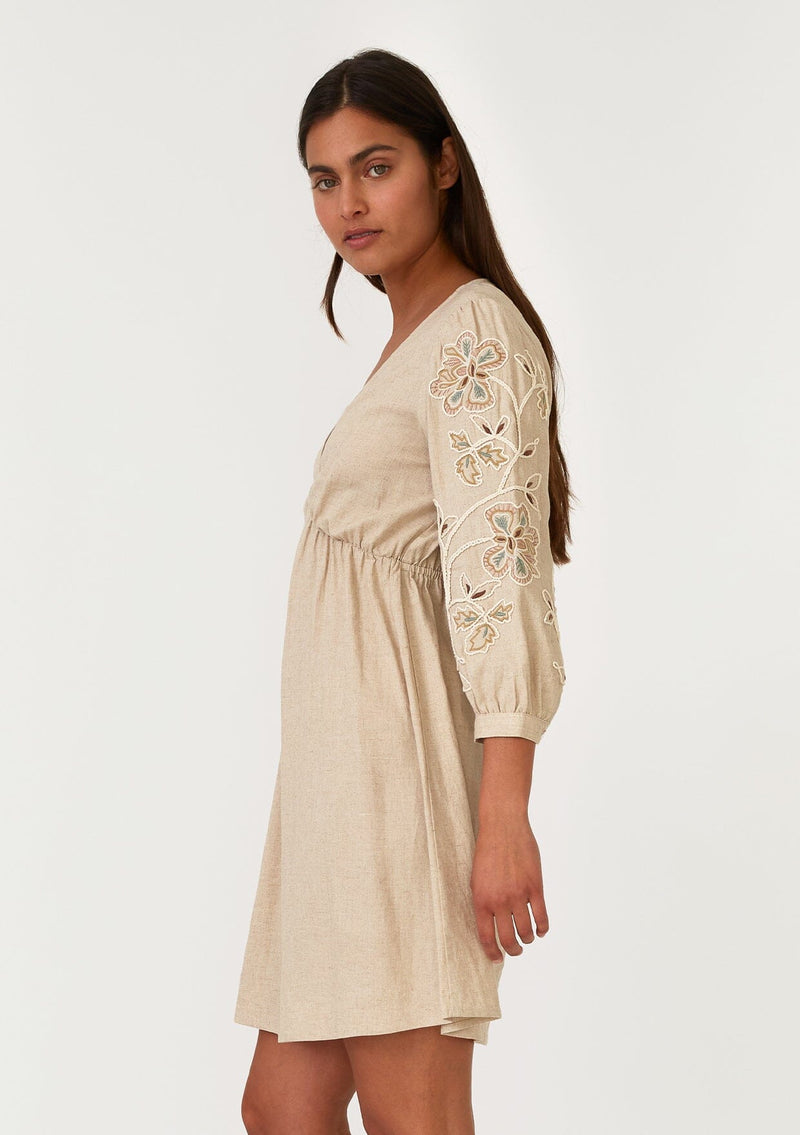 [Color: Natural] A side facing image of a brunette model wearing a classic bohemian natural brown mini dress in a linen blend. With three quarter length sleeves, multi colored embroidered detail, a v neckline, an empire waist, and a relaxed fit. 
