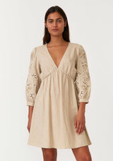 [Color: Natural] A front facing image of a brunette model wearing a classic bohemian natural brown mini dress in a linen blend. With three quarter length sleeves, multi colored embroidered detail, a v neckline, an empire waist, and a relaxed fit. 