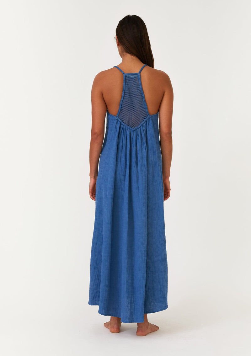[Color: Ocean] A full body back facing image of a brunette model wearing a blue bohemian sleeveless maxi dress crafted from cotton gauze. With braided straps, a v neckline, a relaxed, flowy body, and a sheer crochet mesh racerback detail. 