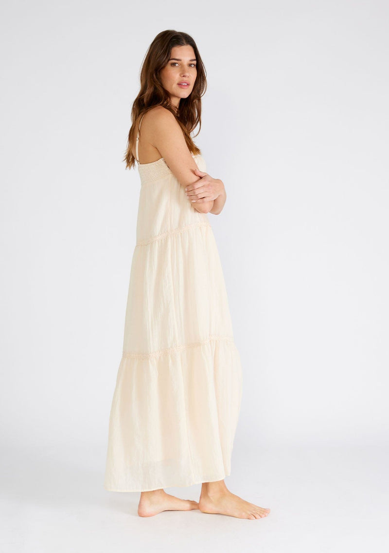 [Color: Light Peach] A side facing image of a brunette model wearing a light peach pink sleeveless maxi dress. A lightweight bohemian style with embroidered detail at the neckline, adjustable spaghetti straps, a lace trimmed tiered skirt, a straight neckline, and a long flowy fit. 