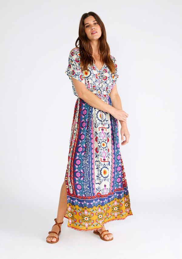 [Color: Natural/Mustard] A front facing image of a brunette model wearing a brightly colorful bohemian maxi dress in a mixed floral print. With short dolman sleeves, a v neckline, a self covered button front top, an adjustable drawstring tie waist, and a long flowy skirt with side slits. 