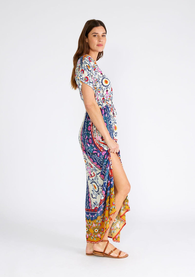 [Color: Natural/Mustard] A side facing image of a brunette model wearing a brightly colorful bohemian maxi dress in a mixed floral print. With short dolman sleeves, a v neckline, a self covered button front top, an adjustable drawstring tie waist, and a long flowy skirt with side slits. 