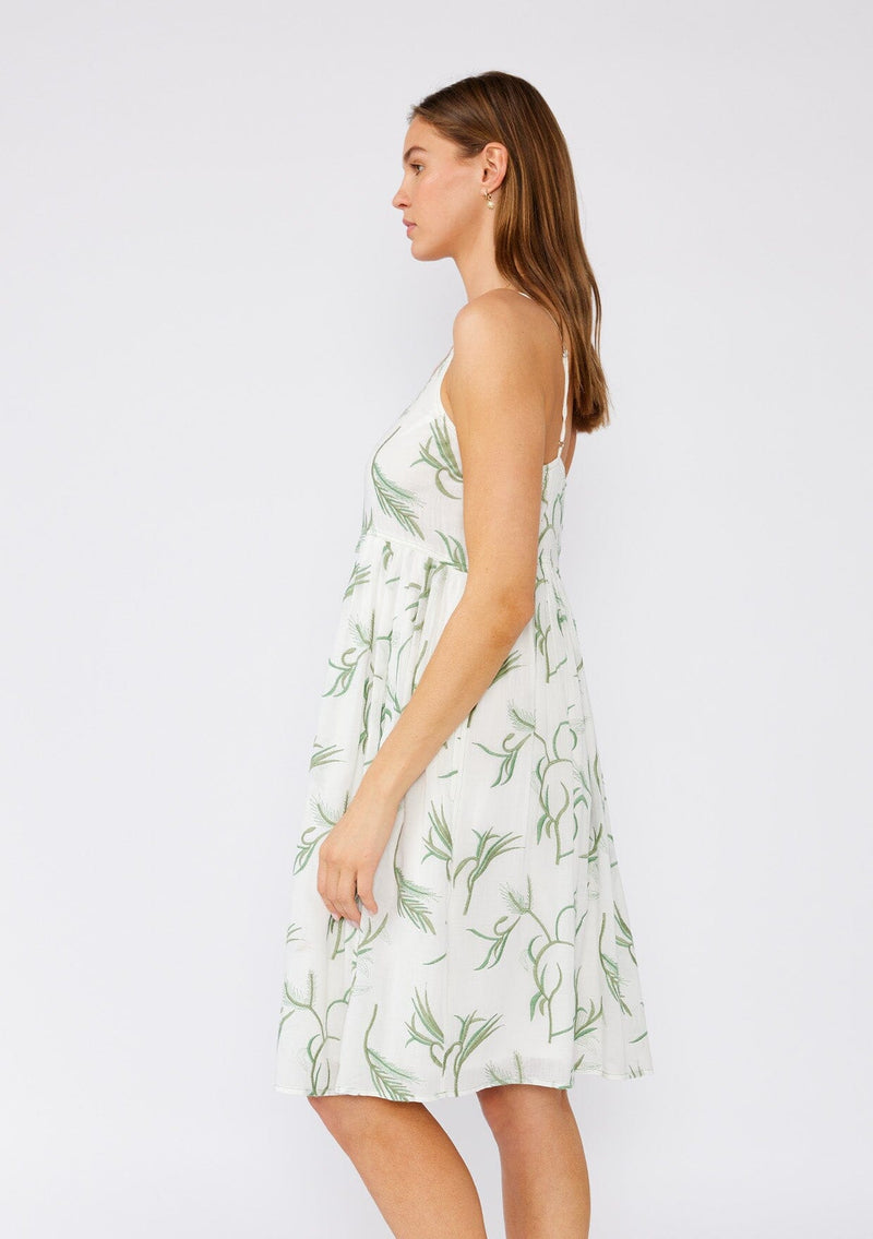 [Color: Off White/Green] A side facing image of a brunette model wearing a sleeveless cotton bohemian mini dress designed in a white and green embroidery. With spaghetti straps, an empire waist, a scooped neckline, side pockets, and a flowy silhouette. 