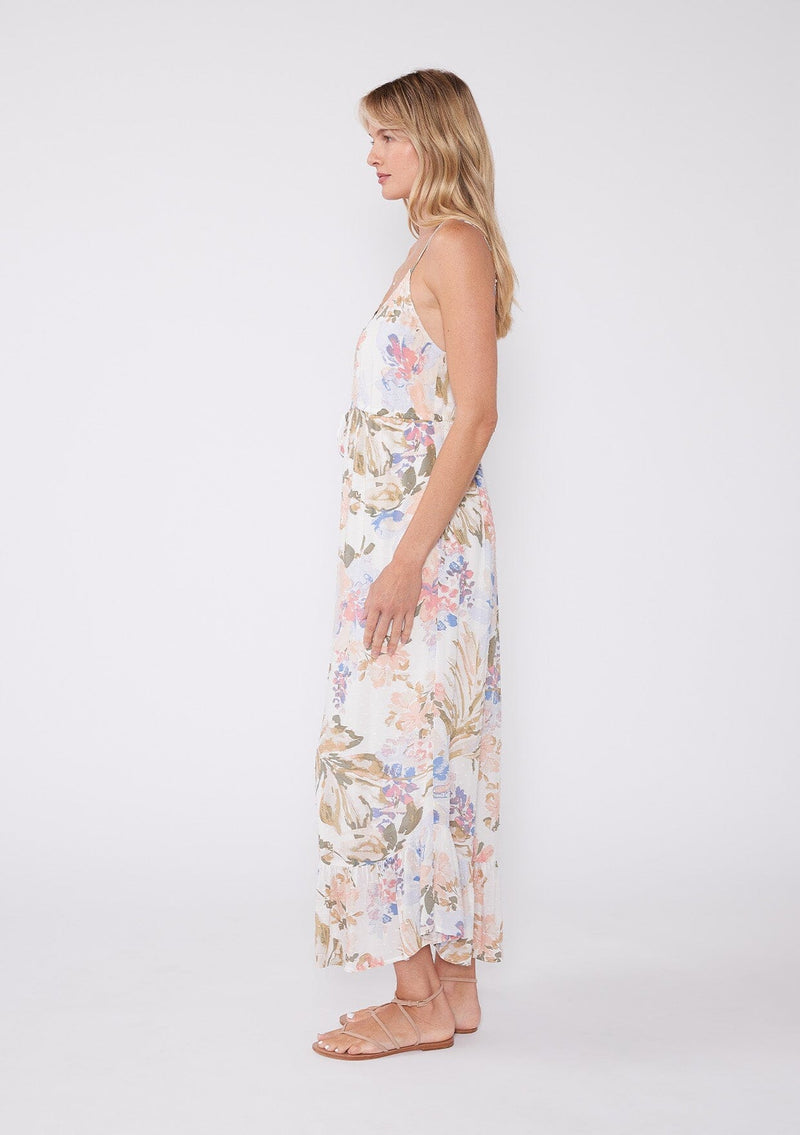 [Color: Off White/Periwinkle] A side facing image of a blonde model wearing a dreamy bohemian mid length sleeveless dress in an off white and periwinkle blue floral print. With adjustable spaghetti straps, a v neckline, a tiered flowy skirt, a button front, and textured clip dot details. 