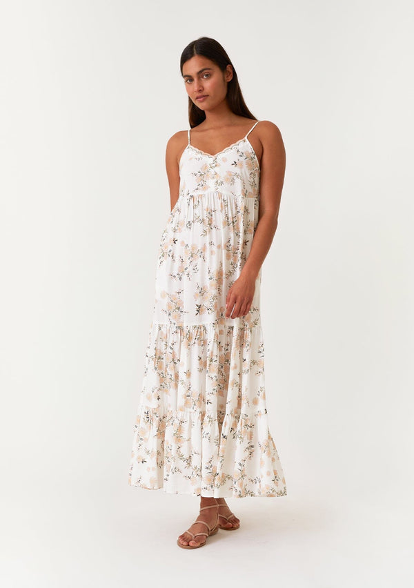 [Color: Ivory/Dusty Coral] A front facing image of a brunette model wearing a pretty bohemian sleeveless maxi dress in a pink and white floral print. With adjustable spaghetti straps, a scalloped trimmed v neckline, a decorative self covered button front top, a tiered flowy skirt, and an empire waist. 