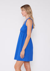 [Color: Cobalt] A side facing image of a blonde model wearing a bright blue sleeveless summer mini dress. With a v neckline, an empire waist, and a back keyhole detail with single button closure.  