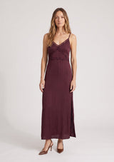 [Color: Merlot] A front facing image of a blonde model wearing a burgundy classic lace trim maxi dress with a v neckline, spaghetti straps, and side slits. 