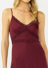 [Color: Merlot] A detail image of a blonde model wearing a burgundy classic lace trim maxi dress with a v neckline, spaghetti straps, and side slits. 