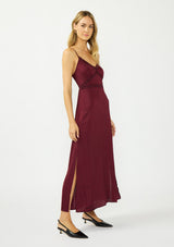 [Color: Merlot] A side facing image of a blonde model wearing a burgundy classic lace trim maxi dress with a v neckline, spaghetti straps, and side slits. 